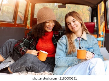 Two Happy Multiracial Girlfriends On Car Trunk In Fall Forest, Embracing And Drinking Hot Tea During Adventure Trip In Autumn Nature, Smiling Female Friends Enjoying Life While Traveling Together