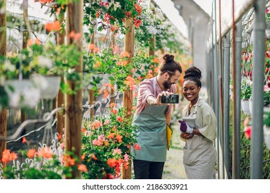 Two Happy Multiracial People Working With Flowers At Industrial Greenhouse While Taking Selfie With A Smartphone. Lifestyle, Gardening, Profession And People Concept. Diverse Couple Working Together.