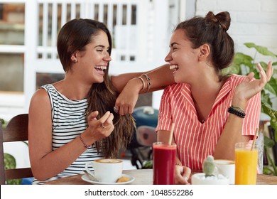 Two Happy Overjoyed Women Sit At Outdoor Cafe, Drink Cocktails, Cappuccino, Tell Funny Stories To Each Other, Being In Good Mood, Laugh Joyfully. Best Female Friends Have Fun Together In Coffee Shop