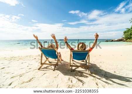 Two happy people having fun on the beach, sitting on blue sunbed with hands raised up, spending leisure time together. Summer holidays concept. Tourism. Travelers.
