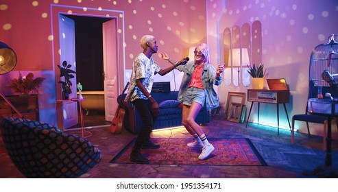 Two happy positive multi-ethnic youth dancing moving rhythmically in good mood at home party, male and female friends having fun energetically moving in room in neon disco light, retro style concept