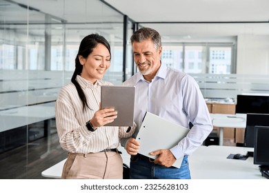 Two happy professional business people team Asian woman and Latin man workers working using digital tablet tech discussing financial market data standing at corporate office meeting. - Shutterstock ID 2325628103