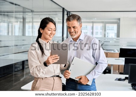 Two happy professional business people team Asian woman and Latin man workers working using digital tablet tech discussing financial market data standing at corporate office meeting.