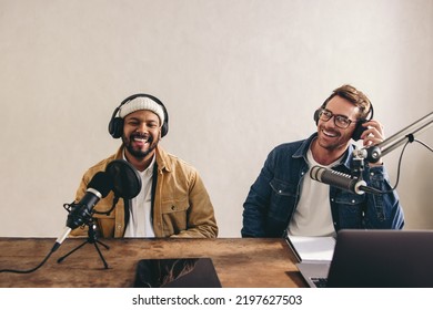 Two happy radio presenters having a good time on air. Young men smiling happily while recording an audio broadcast in a studio. Cheerful content creators co-hosting an internet podcast.