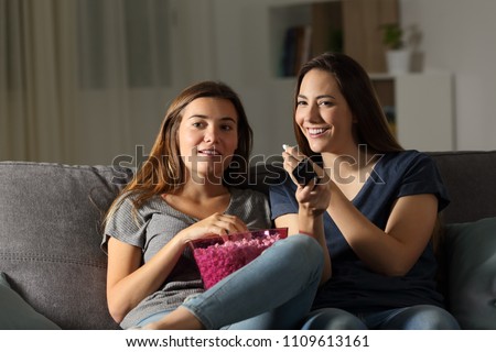 Two happy roommates wathing tv in the night sitting on a couch in the living room at home