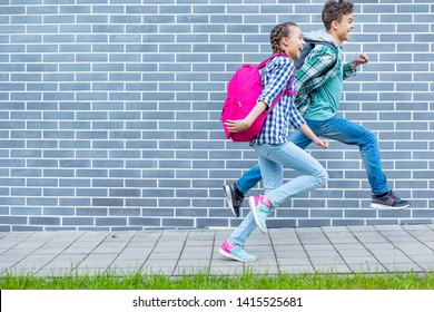 Two Happy Schoolchildren With Backpacks Run To School On Street Next To An Brick Wall. Cheerful Cute Children Pupils Teen Girl And Boy Back To School. Concepts Of Friends, Childhood And Education.