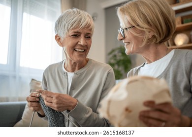 Two happy senior women enjoying their free time, knitting together and chatting in the living room, sharing a moment of relaxation and friendship