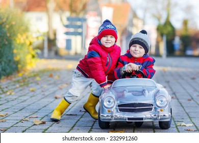 Two Happy Sibling Boys In Red Jackets And Rain Boots Playing With Big Old Toy Car, Outdoors.  Kids Leisure On Cold Day In Winter, Autumn Or Spring.