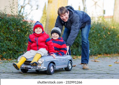 Two Happy Sibling Boys In Red Jackets And Rain Boots And Their Father Playing With Big Old Toy Car, Outdoors. Family And Kids Leisure On Cold Day In Winter, Autumn Or Spring.