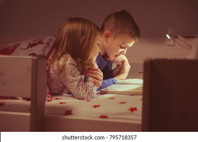 Two happy sibling children reading book in bunk bed under blanket
