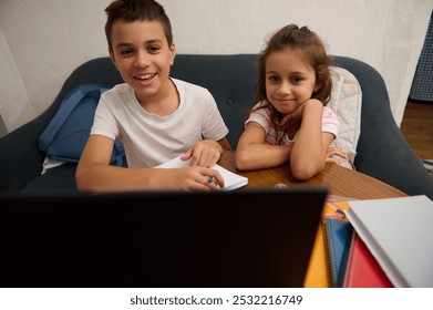 Two happy siblings sitting at a table, engaged in online learning. They appear joyful and attentive, surrounded by school supplies. - Powered by Shutterstock