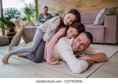 Two Happy Sisters Having Fun With Father, Lying On His Back With Mother At Background At Home