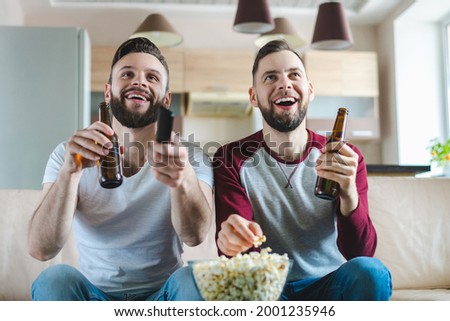 Two happy smiling buddies sitting on sofa, watching football play live broadcast and celebrating their favourite team victory.