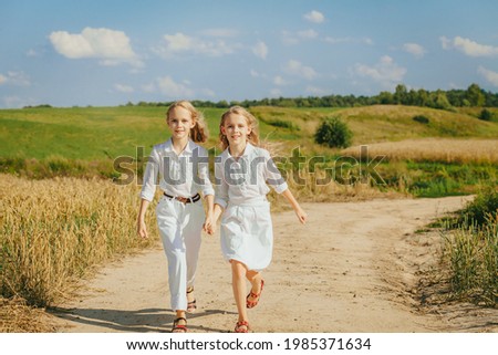 Two happy smiling girls walk in countryside road.  Outdoor photo of blonde twins sisters posing on nature background. Summer vacation in countryside. 