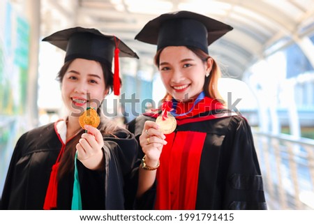 Two happy smiling graduated students hold gold medal award, young beautiful Asian women look at camera on commencement day, celebrating successful education on graduation day from university.