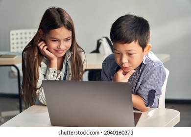 Two happy smiling interested preteen diverse schoolmates working together watching learning video using laptop computer during lesson in classroom. Modern technologies for education concept.