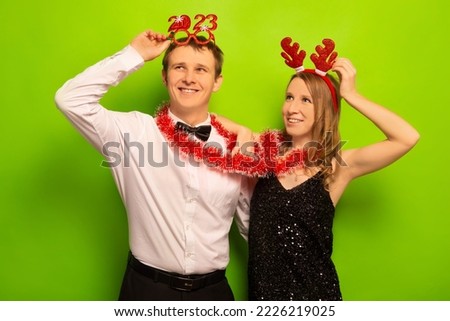 Two happy smiling people husband and wife or friends in funny party glasses, Christmas reindeer antlers headwear with tinsel looking up on a green background. New Year 2023 and Xmas celebration.
