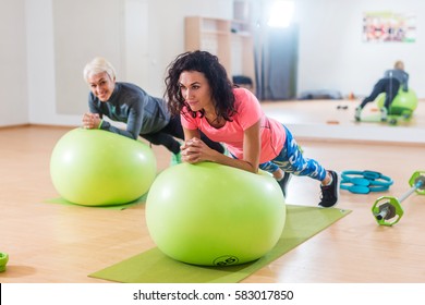 Two Happy Sportswomen Exercising With A Swiss Ball Doing Plank Exercise In Gym.