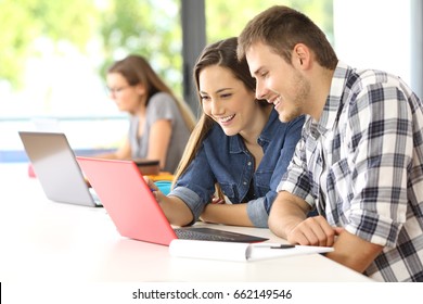Two happy students learning on line together with a computer in a classroom with other classmate in the background