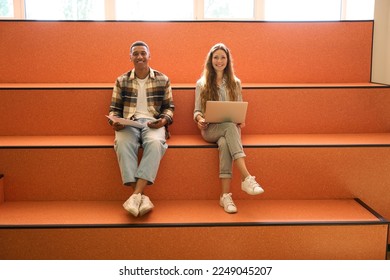 Two happy students sitting and using gadgets in university