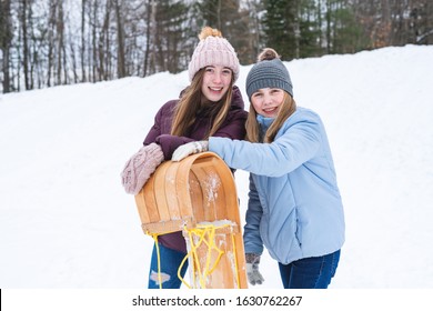 Two Happy Teen Girls/friends Standing Behind A Wood Sled/toboggan While Standing At The Bottom Of Hill Covered With Snow.