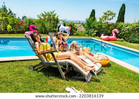 Two happy teenage girls sit near the pool on chaise longue in the garden chilling