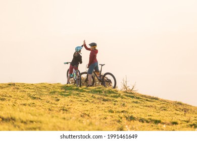 Two Happy Woman High Five Over The Sunset After A Successful Mountain Biking Trip In The Mountains. Celebrate A Cross Country Cycling Journey.