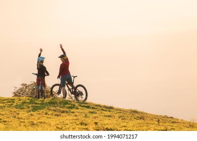 Two Happy Woman High Five Over The Sunset After A Successful Mountain Biking Trip In The Mountains. Celebrate A Cross Country Cycling Journey.