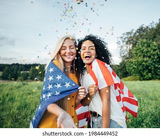 Two Happy Women Covered By The USA Flag. Young Friends Celebrating The 4th Of July Throwing Confetti On A Field.