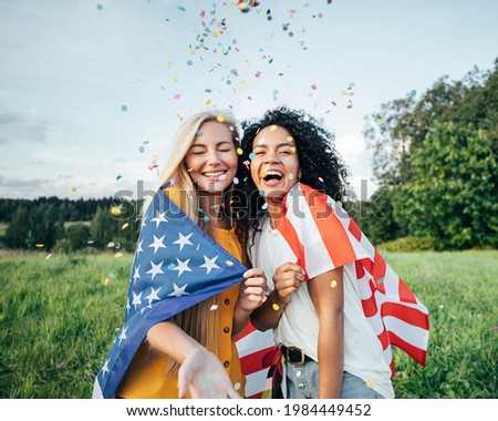Two happy women covered by the USA flag. Young friends celebrating the 4th of July throwing confetti on a field.
