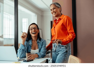 Two happy women employees laughing and collaborating in a colorful and modern office setting, enjoying coffee during a meeting. - Powered by Shutterstock