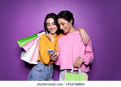 Two happy women friends shoppers holding shopping bags using mobile apps for online shopping standing on purple background. Retail ecommerce fashion sale offers, mall discounts in applications concept