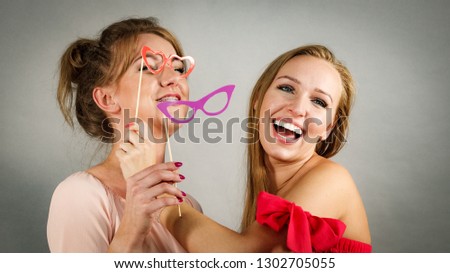 Two happy women holding symbols on stick having fun pretending to wear eyeglasses. Photo and carnival funny accessories concept.