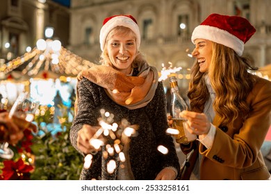 Two happy women in Santa hats enjoying Christmas market festivities. Friends hold sparklers and drinks, laughing together amid twinkling lights, decorations - Authentic holiday spirit and friendship. - Powered by Shutterstock