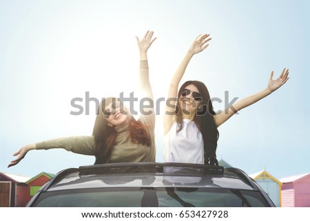 Two happy women wearing sunglasses, enjoying freedom on car sunroof near the cottage 