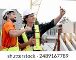 Two happy worker engineers with safety vest and helmet use mobile phone to take a selfie with coworker at construction building Architect labors work and take photo together at building site workplace