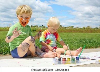 Two Happy Young Children, A Little Boy And His Baby Brother, Are Sitting Outside On A Summer Day, Painting A Picture, And Covering Themselves In Paint.