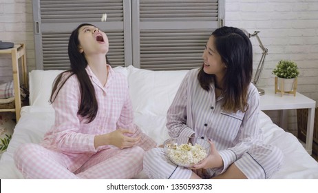 Two Happy Young Female Asian Friends Sitting On Bed And Eating Popcorn From Glass Bowl. Funny Girl In Pink Pajama Throwing Food Using Mouth Catching. Best Sisters Enjoy Relax Leisure Time In Bedroom.