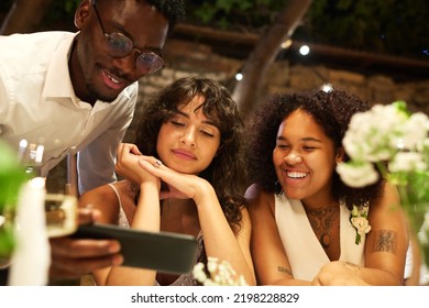 Two Happy Young Lesbian Brides And Black Guy Communicating In Video Chat While Sitting By Served Festive Table At Wedding Party