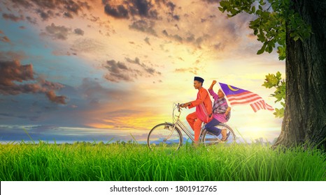 Two happy young local boy riding old bicycle at paddy field holding a Malaysian flag with beautiful sunset moments. Independence Day concept