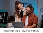 Two happy young programmers looking at laptop screen with coded data and consulting about ways of decoding it at meeting in dark office