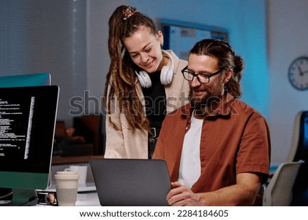 Two happy young programmers looking at laptop screen with coded data and consulting about ways of decoding it at meeting in dark office