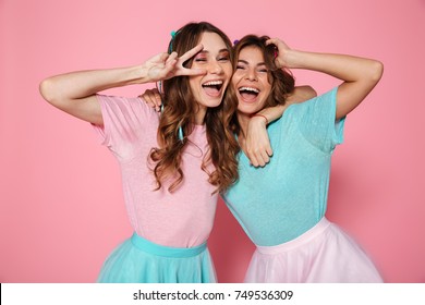 Two happy young woman in colorful clothes showing peace gesture, looking at camera, isolated over pink background