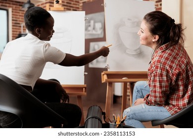 Two happy young women friends learning new things together, attending group art class, discussing artwork and smiling. Young diverse people talking communicating during drawing class for adults - Powered by Shutterstock