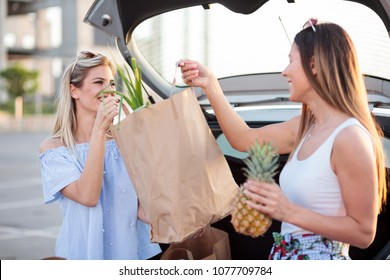 Two Happy Young Women Loading Paper Grocery Bags Into A Car Trunk.