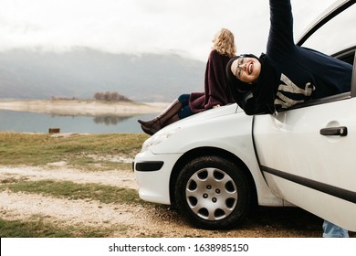 Two happy young women on car trip. They are having fun while one is sitting on the car and one with hijab is near window. They are smiling and enjoying their time together.