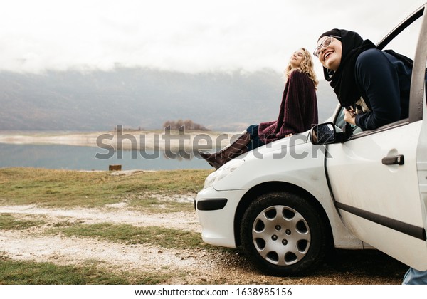 Two happy young\
women on car trip. They are having fun while one is sitting on the\
car and one with hijab is near window. They are smiling and\
enjoying their time\
together.