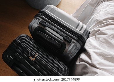 Two hard shell suitcases rest. Black luggage stands beside gray. Closed zippers secure bags. Cases lean against bedding. Travel preparation background suits vacations. Wooden floor surrounds bags.
