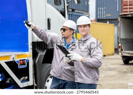 Two hardhat clad logistics workers inspect a truck beside stacked shipping containers, using a clipboard while pointing during loading operations.
