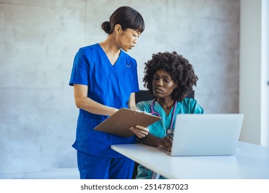 Two healthcare workers in scrubs discuss patient notes while using a laptop. Teamwork and communication in a medical setting are emphasized. - Powered by Shutterstock